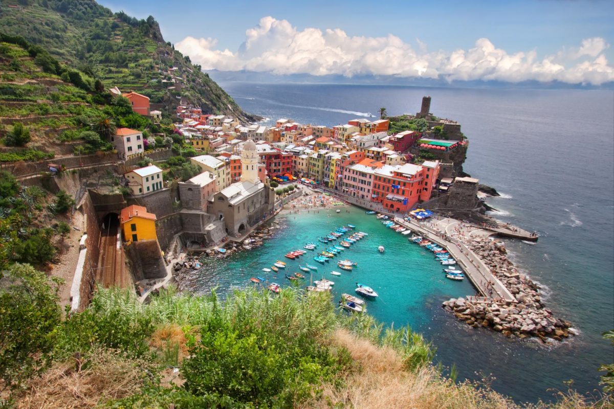 This colorful little town by the sea in Italy. In Vernazza, which is one of the five famous villages of Cinque Terre, you can see bright buildings stacked on hills next to the ocean. Small boats float in the harbor, and people walk along narrow streets to shops and cafés. The water is blue and sparkling, and the whole town looks like it belongs in a postcard.
