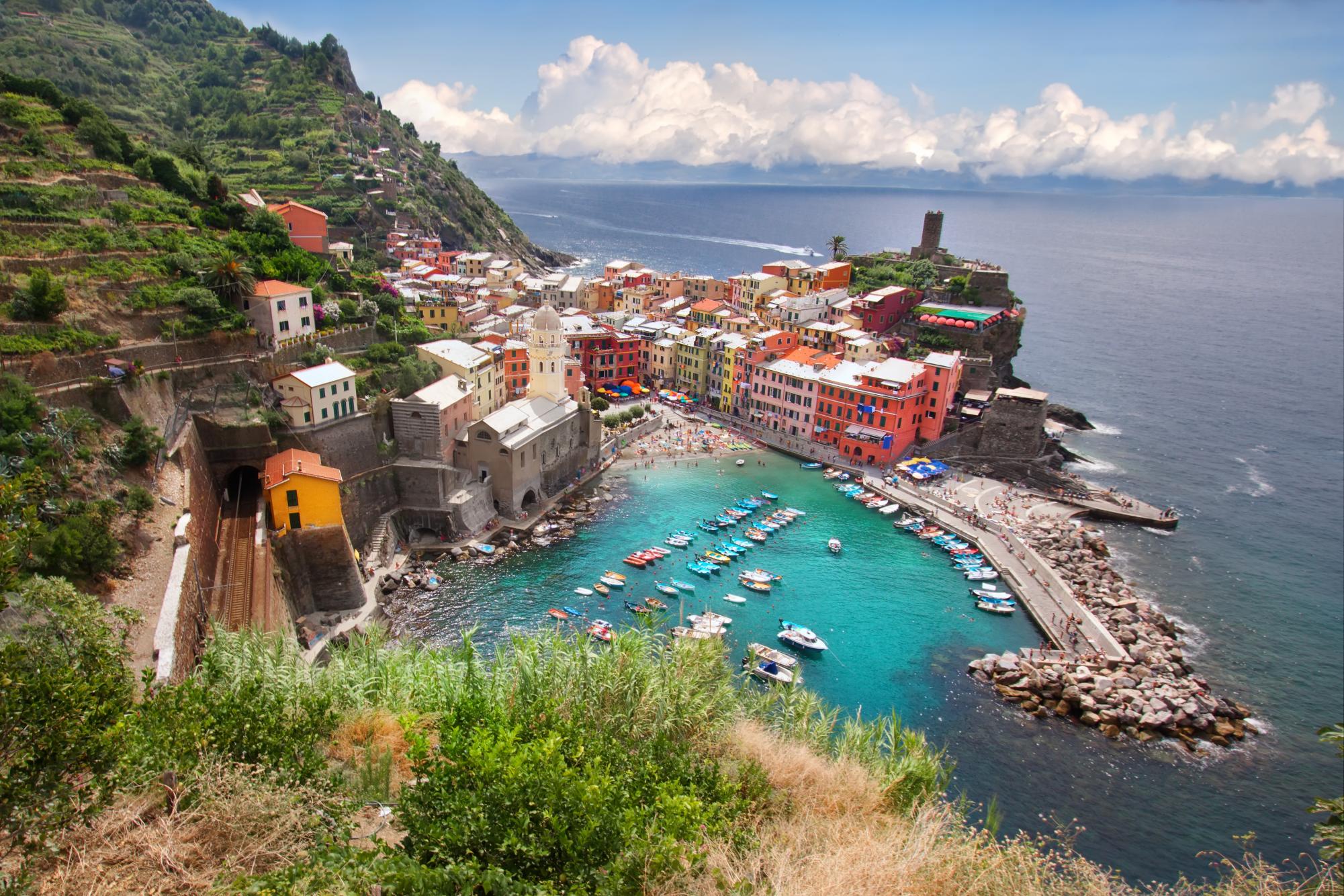 This colorful little town by the sea in Italy. In Vernazza, which is one of the five famous villages of Cinque Terre, you can see bright buildings stacked on hills next to the ocean. Small boats float in the harbor, and people walk along narrow streets to shops and cafés. The water is blue and sparkling, and the whole town looks like it belongs in a postcard.